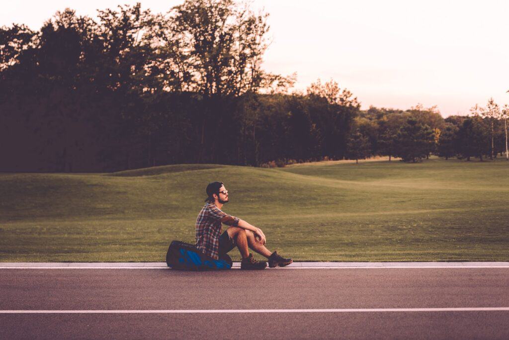 tired-traveler-side-view-of-handsome-young-man-sitting-on-his-backpack-at-the-side-of-the-road-and-looking-thoughtful