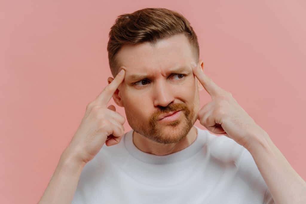 close up shot of serious bearded adult man keeps fingers on temples concentrated aside tries to recall something in mind dressed casually isolated over pink background male model thinks deeply
