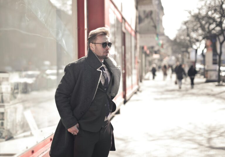 A fashionable man in a coat and sunglasses walking on a sunny city street